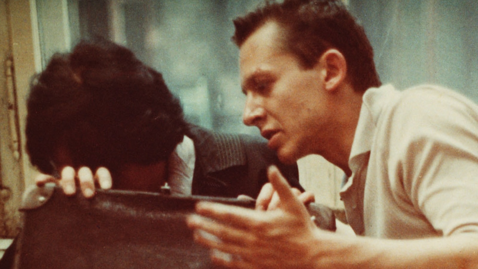 A man gestures aggressively at a woman, who hides her head behind her bag as they sit in public.