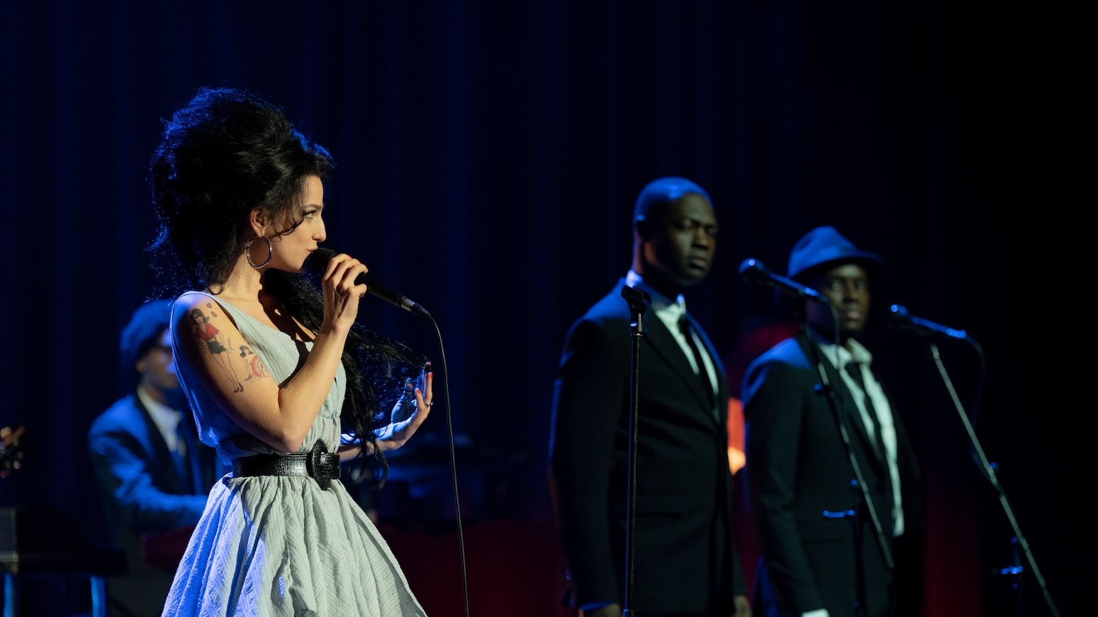 A woman dressed as singer Amy Winehouse, in an upswept black hairdo, holds a microphone on a stage, while two men in black suite slightly out of focus are on the right side of the stage
