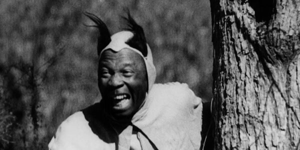 A medium brown skinned man in a devil costume with horns peers from behind a tree with his mouth open in a diabolical laugh