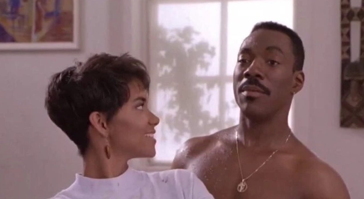 A short-haired woman in a pink-colored sweatshirt and a shirtless man wearing a gol necklace standing close behind her as she looks back at him and he looks off-screen talking to someone else