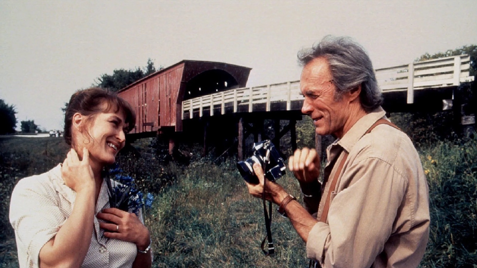 Standing in front of a covered bridge in a rural setting, a man holds a camera, pointing a lens towards a woman who blushes as she grabs her neck.