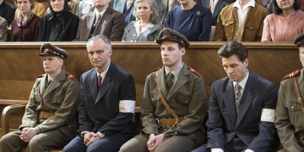 A row of men in a courtroom, who appear to be the defendants on trial, two of them wear blur suits and white armbands, the other two are dressed in military uniforms, all in WWII-era outfits