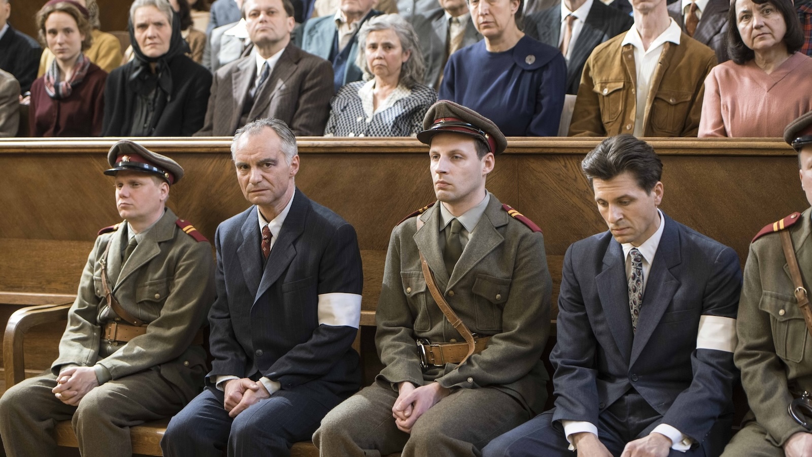Charlatan A row of men in a courtroom, who appear to be the defendants on trial, two of them wear blur suits and white armbands, the other two are dressed in military uniforms, all in WWII-era outfits