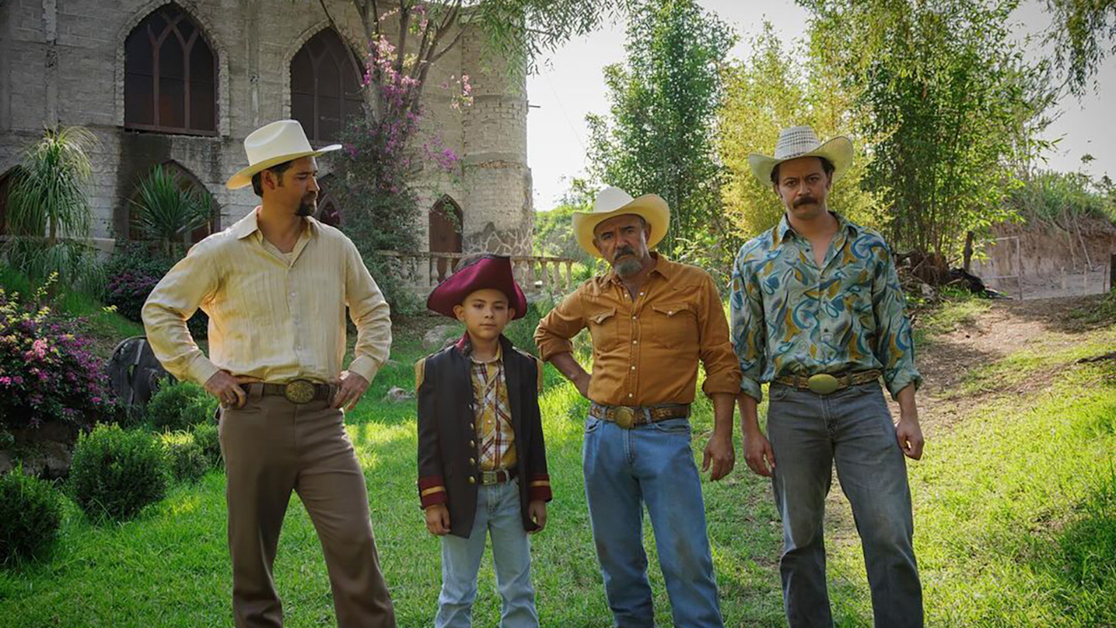 Four people—three men in cowboy hats and a young boy in a tricorne hat—stand in front of an opulent villa surrounded by green trees.