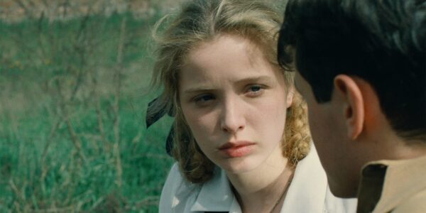 A blonde young woman with braids looks at a man in profile; they are against a green outdoor backdrop