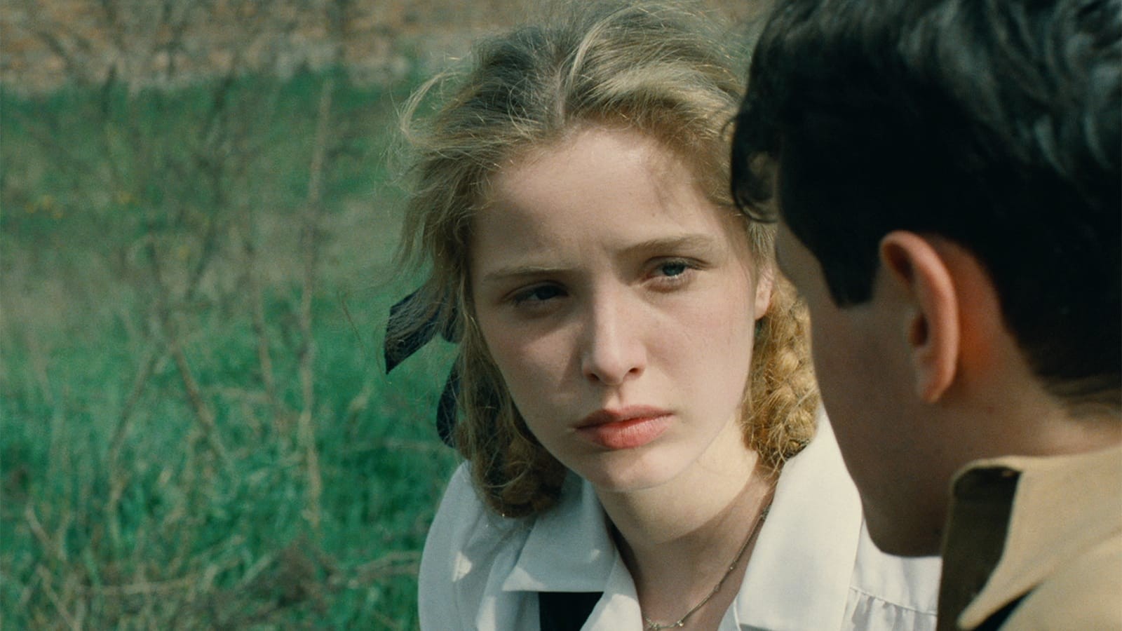 A blonde young woman with braids looks at a man in profile; they are against a green outdoor backdrop