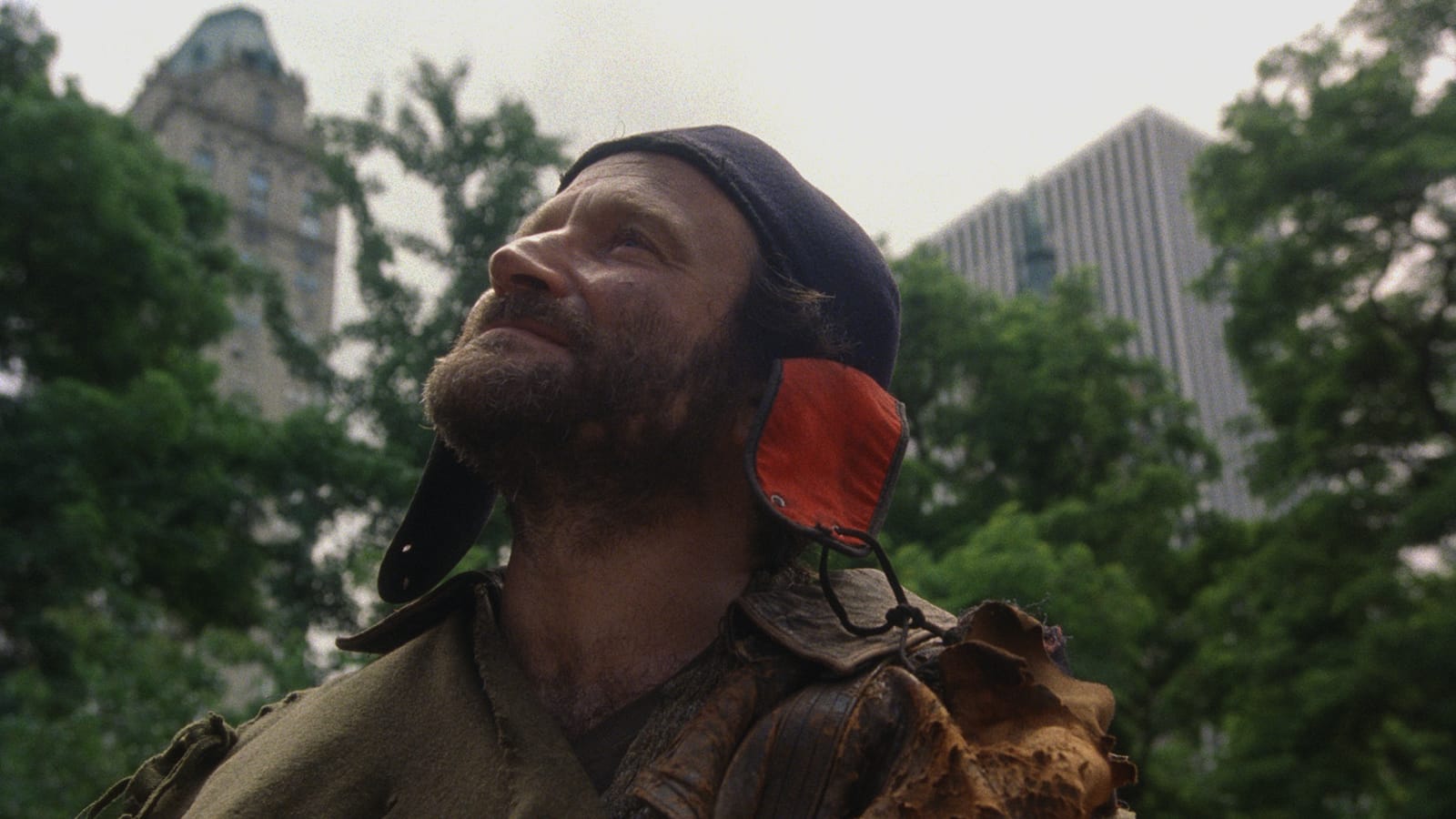 Fisher King A man in a winter hat with ear flaps and a beard looks up in Central Park in New York, surrounded by trees and buildings.