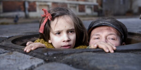 Two children, the girl with a red ribbon in her hair and a boy with a cap, poke their heads out of a sewer grate