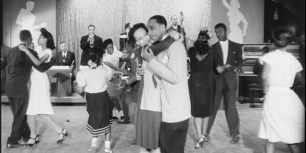An African American woman and man dance together cheek to cheek amidst other dancers at a juke joint in this black and white image from the 1940s.