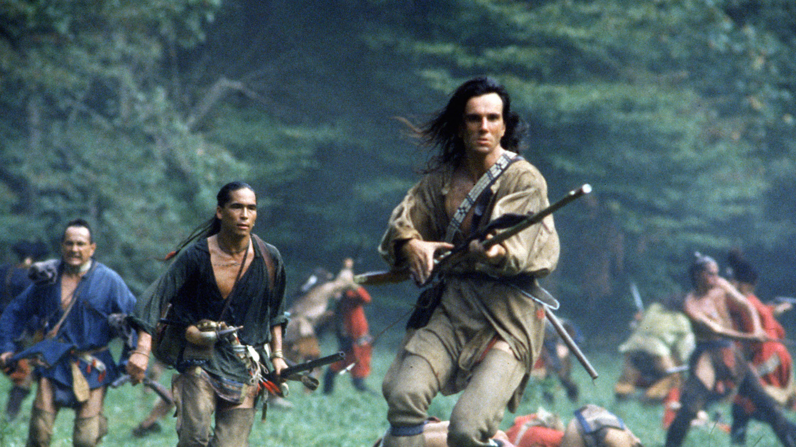 A long-haired man with a rifle, surrounded by other Native American warriors, runs through a field surrounded by trees and mountains