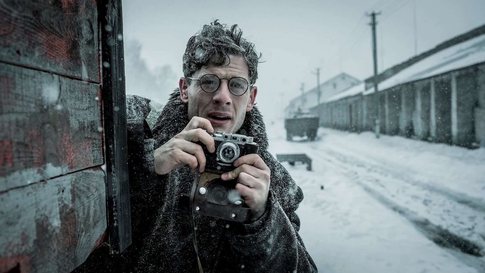 Against a wintry railroad tracks landscape a man holds up a camera and is about to take a photo