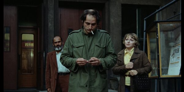 A man in green fatigues lights is about to light a cigarette dangling from his mouth, while a man and woman walk behind him, looking expectantly at him