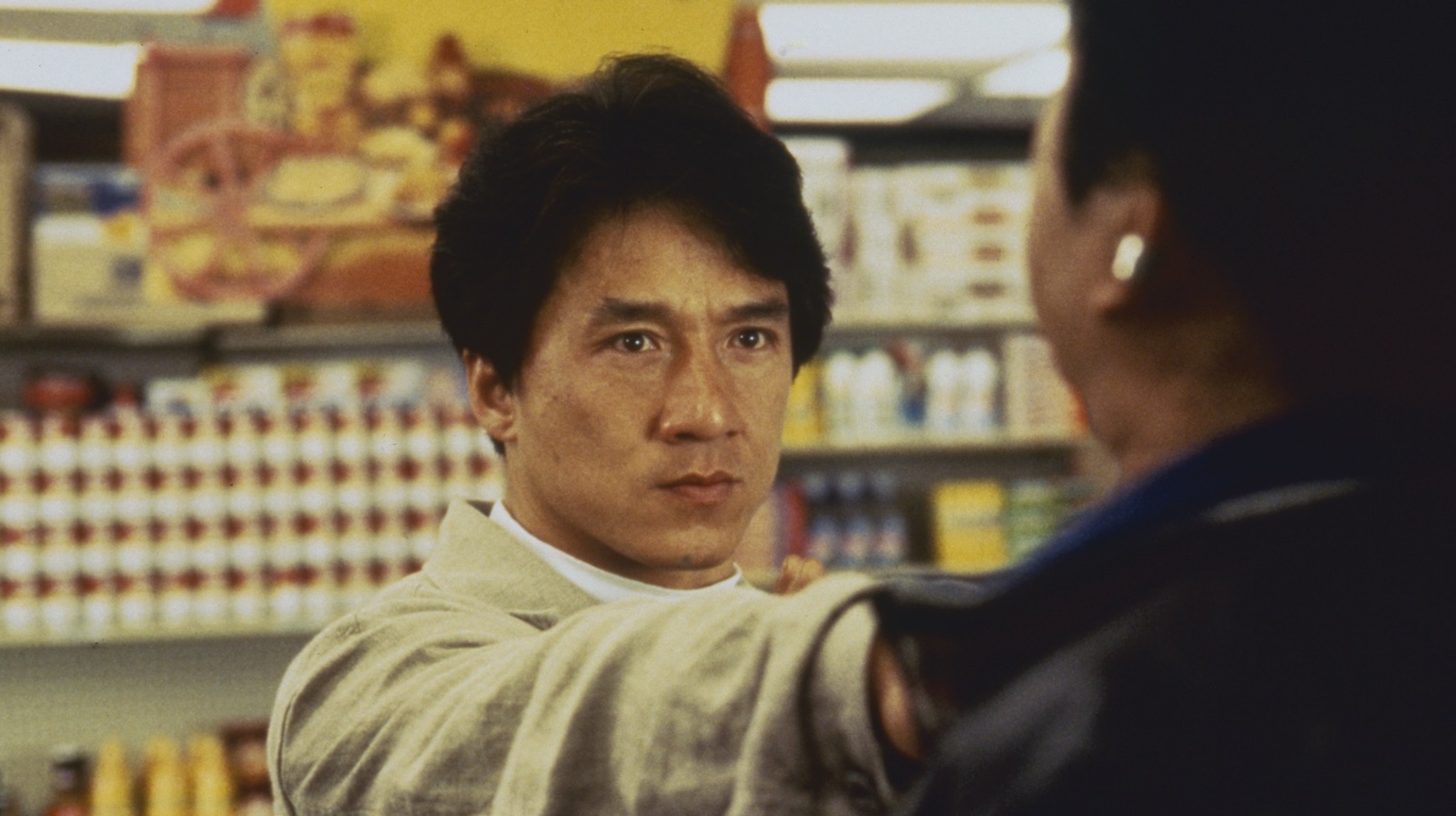 A man stands in a supermarket with a threatening hand thrust in front of him grabbing someone's neck, which we see from behind