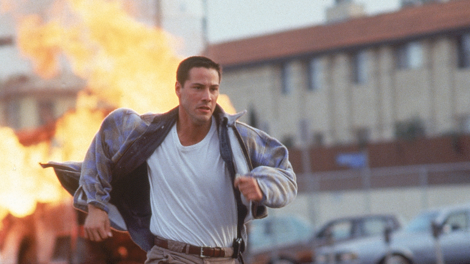 A man wearing a t-shirt and jacket and khakis runs from a blazing explosion in the background