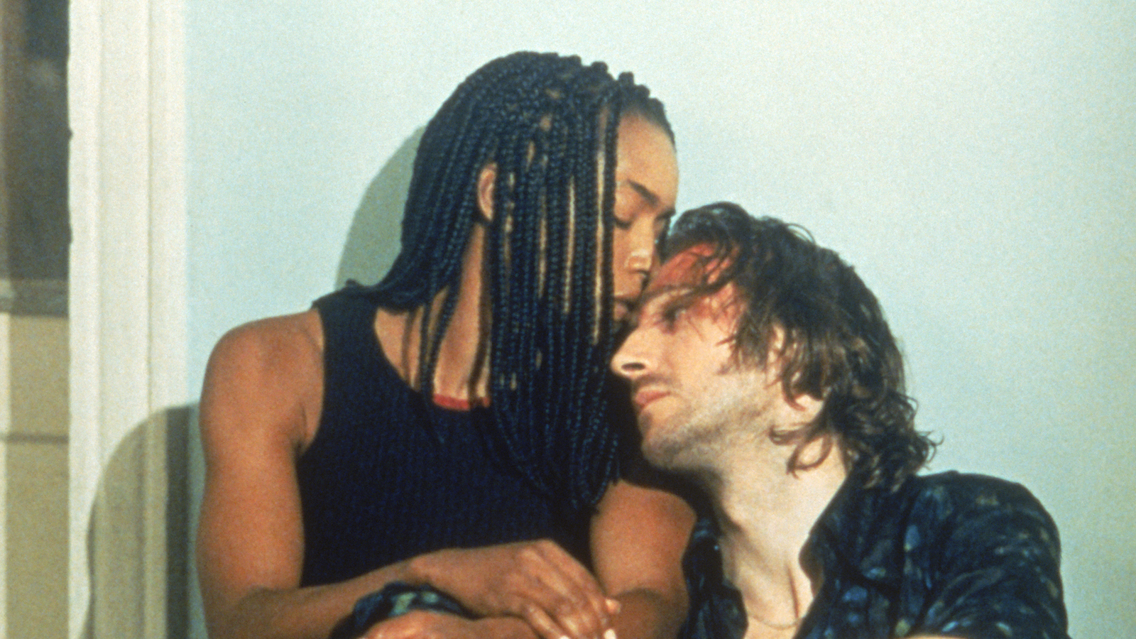 A brown-skinned woman in dreadlocks kisses the forehead of a light-skinned man with a bloody forehead as they crouch against a white wall