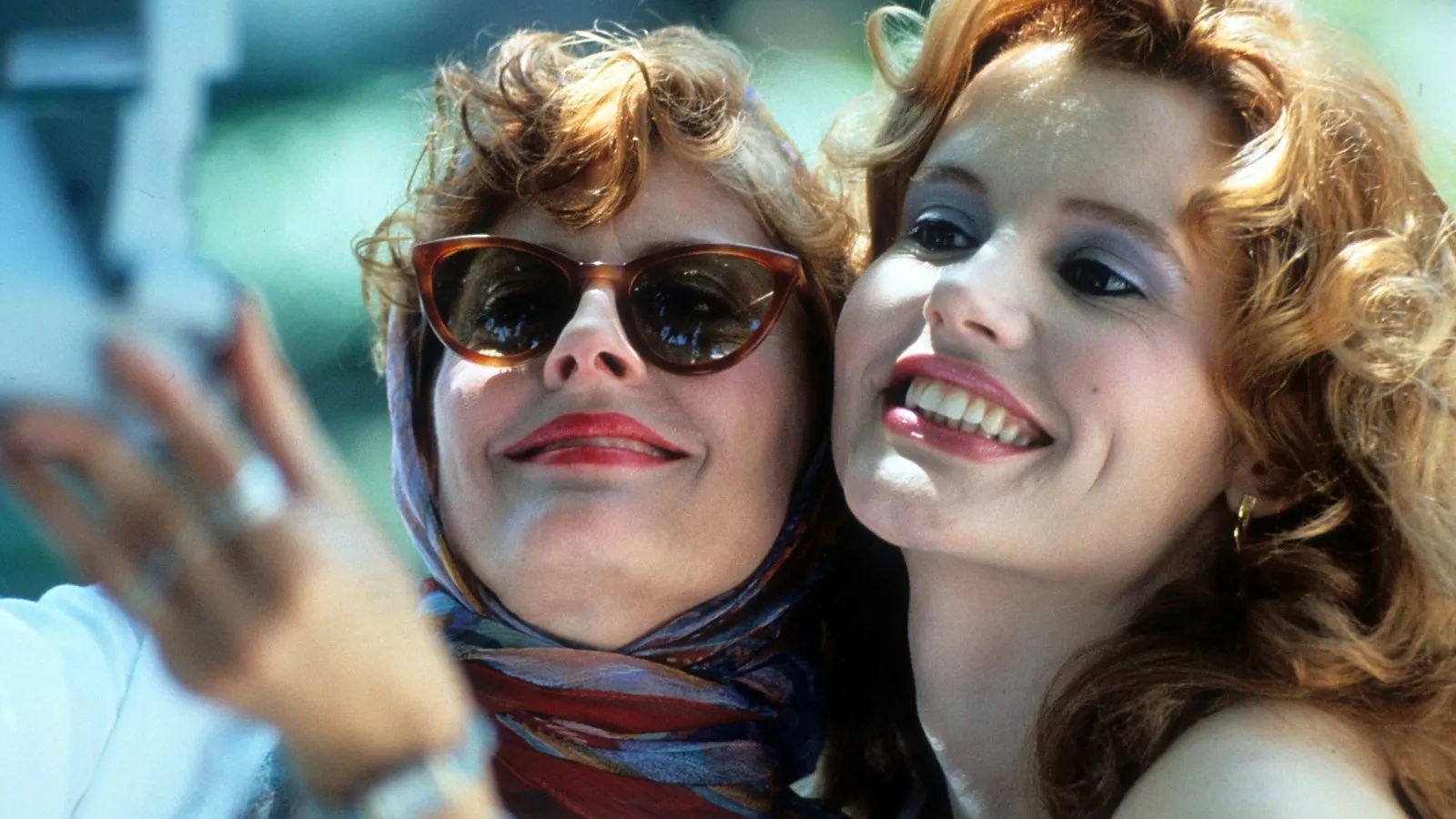 Thelma and Louise Two women smile take a polaroid of themselves, both wearing makeup, the one on the left in sunglasses and kerchief.