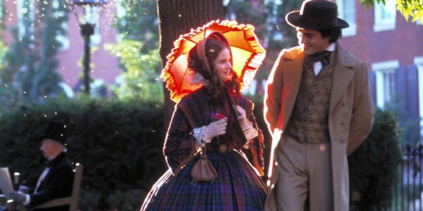 A woman with a yellow and orange parasol lit up by the sun walks down a city street next to a man in a hat; they are smiling and are wearing late nineteenth-century-style clothes