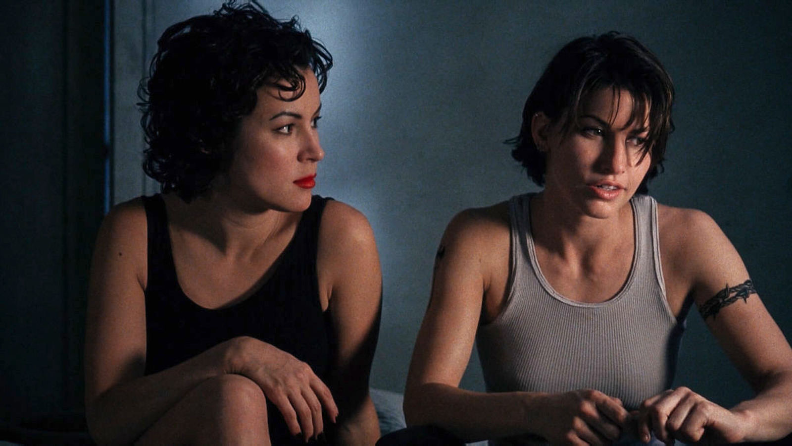 Two women, one traditionally feminine in a black dress and lipstick and the other more butch in a gray tank top sit next to each other in a dark room