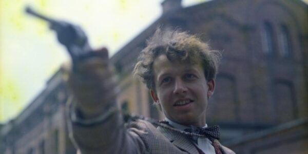 A man raises a pistol to something off camera on a city street, smiling