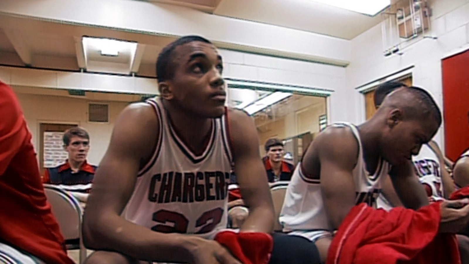 A young man of medium brown skin wearing an athletic tank top with the word CHARGERS on it sits in a locker room and looks pensively.