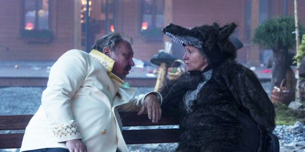 Two people sit on a bench looking at one another: the man is in a white jacket, the woman is wearing a wolf costume