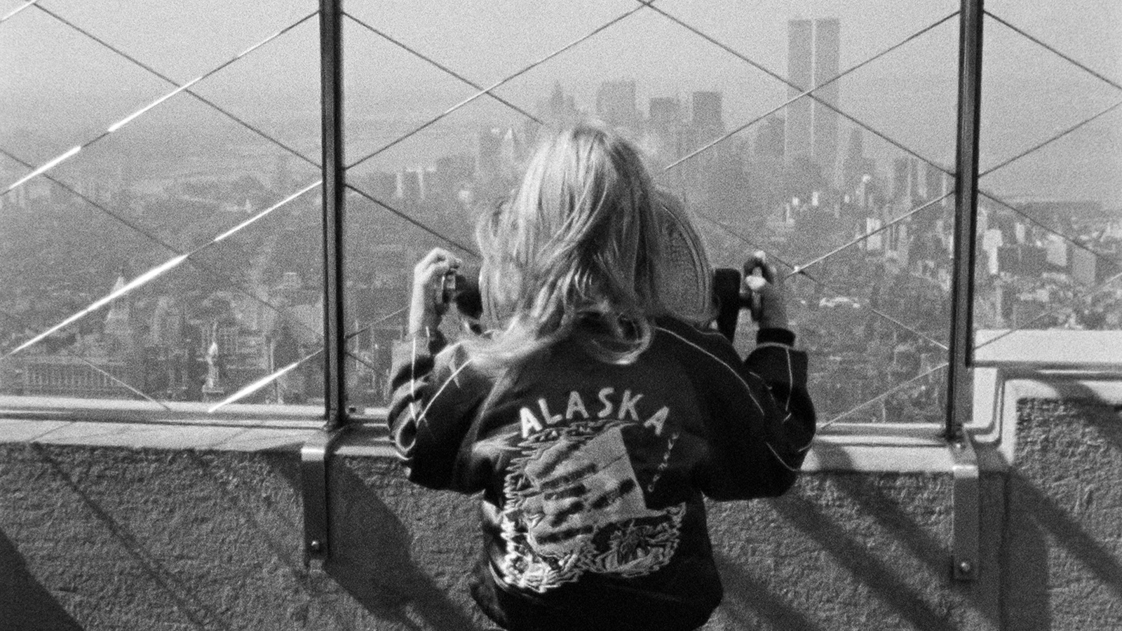 A young child with a jacket reading ALASKA on the back looks out a viewfinder at the top of a skyscraper in New York City, her back to us. The World Trade Towers can be seen in the haze in the distance.