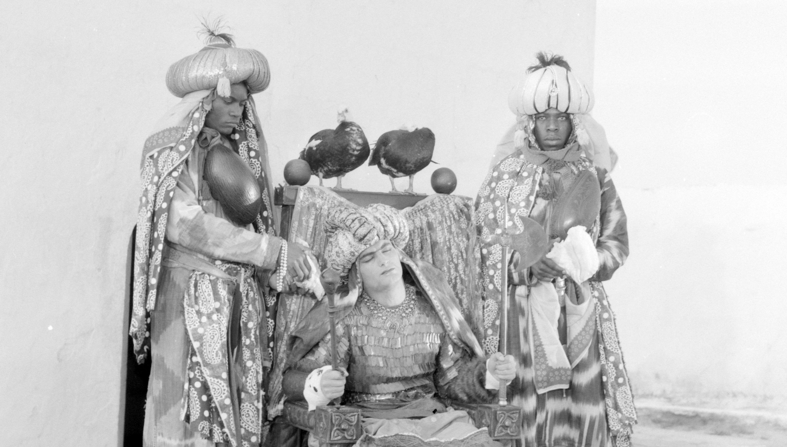 A light skinned sultan in ornate clothes sits on a throne flanked by two darked skinned men standing next to the chair.