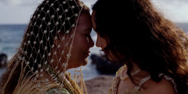 Two women with long hair stand with their foreheads touching against a beach landscape