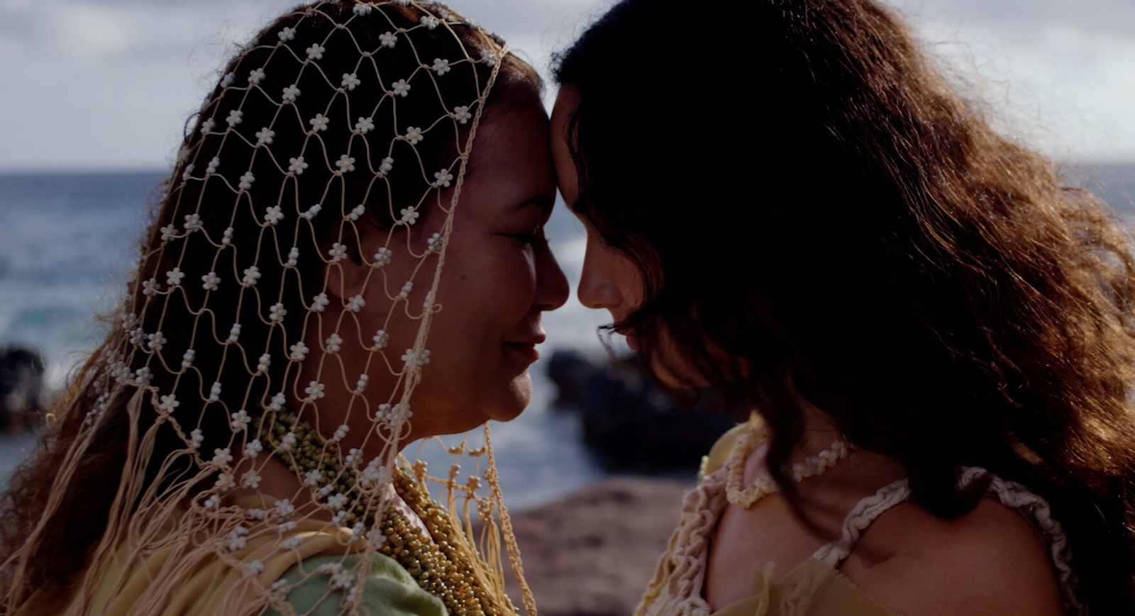 Two women with long hair stand with their foreheads touching against a beach landscape