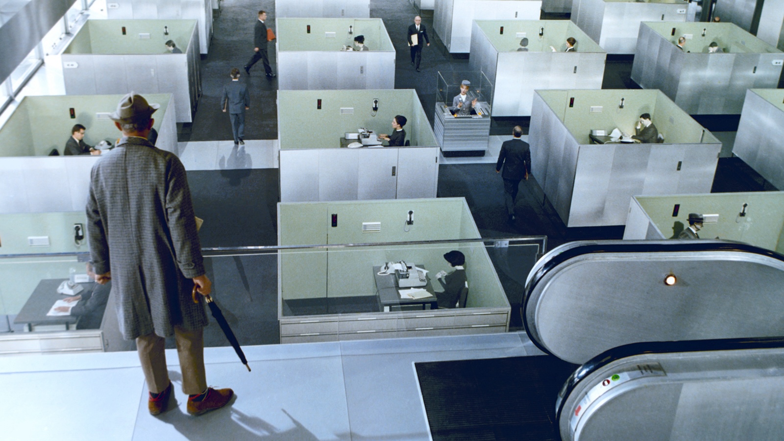 A man, seen from behind, holding a closed umbrella and wearing a trenchcoat and rumpled hat stands near the top of an escalator, looking down on an vast office of cubicles.