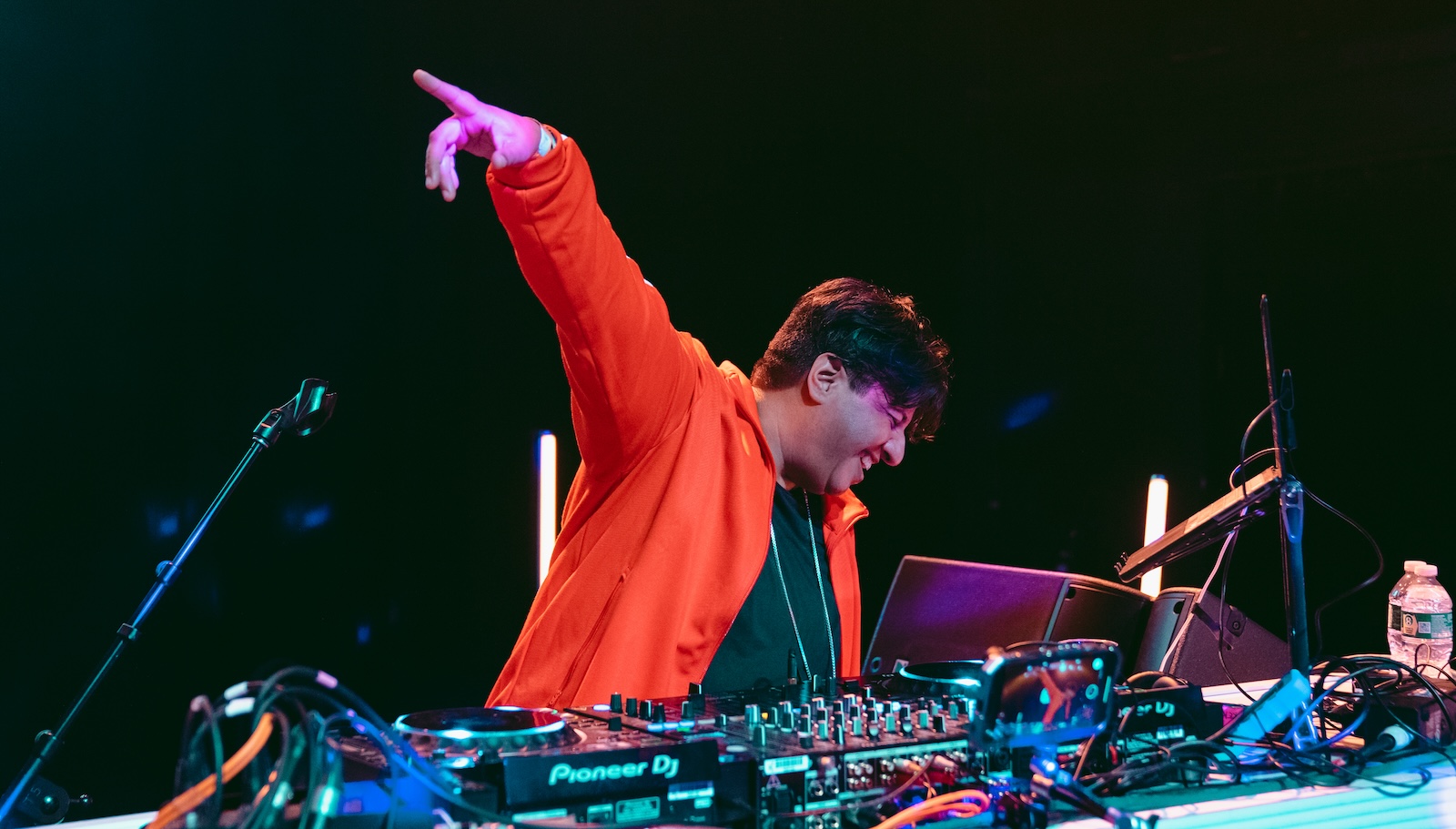 A DJ in a red jacket points to the sky as he plays over his mix table covered in DJ audio equipment