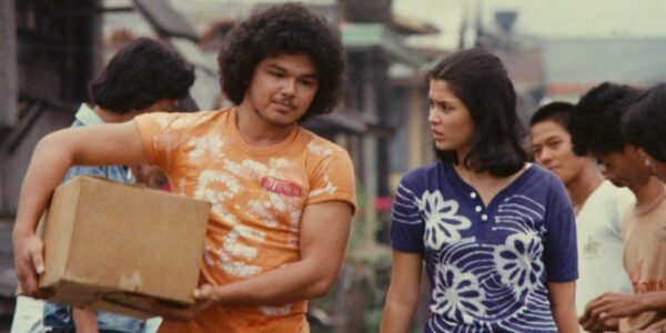 A man and woman in colorful shirts, orange and blue, respectively, walk down a city street; he carries a brown cardboard box under his arm and she looks at him quizzically.