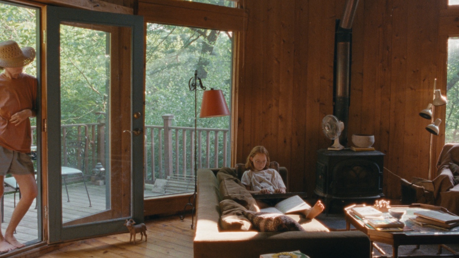 Janet Planet_03 A red headed young girl with glasses sits on a couch in a modern looking living room in the afternoon while a woman stands and watched her from the door on the left.