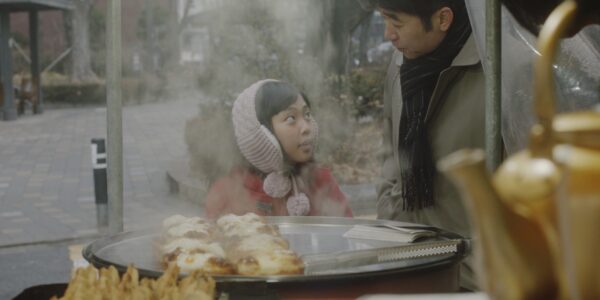 A little girl in a pink winter hat with pom poms looks up at an older man, maybe her father, while standing in front of a food cart with a tray of pastries on it