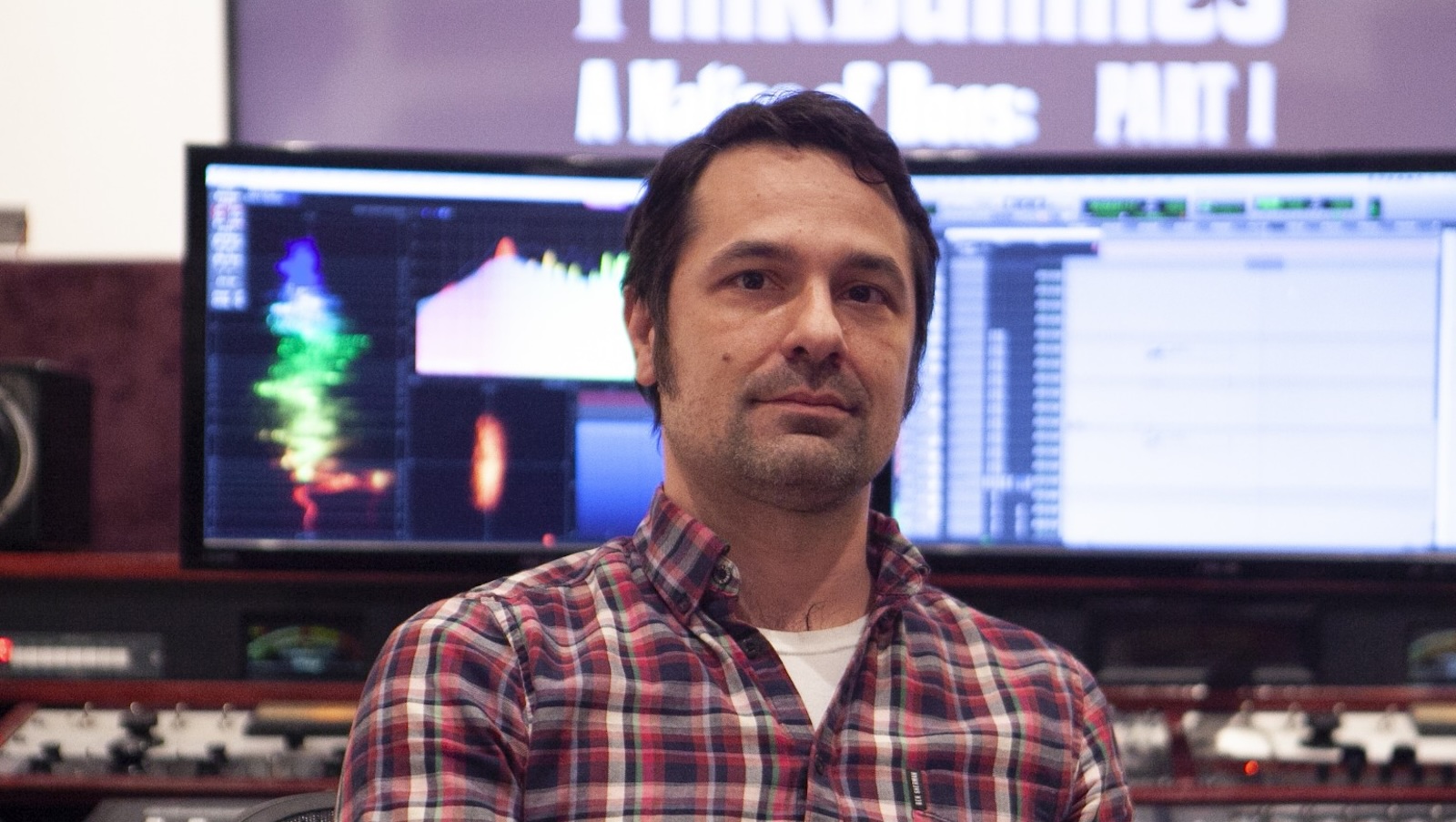 Matt Rocker A dark-haired man wearing flannel shirt and a serious expression looks directly at us while sitting in front of two large computer monitors displaying film editing software