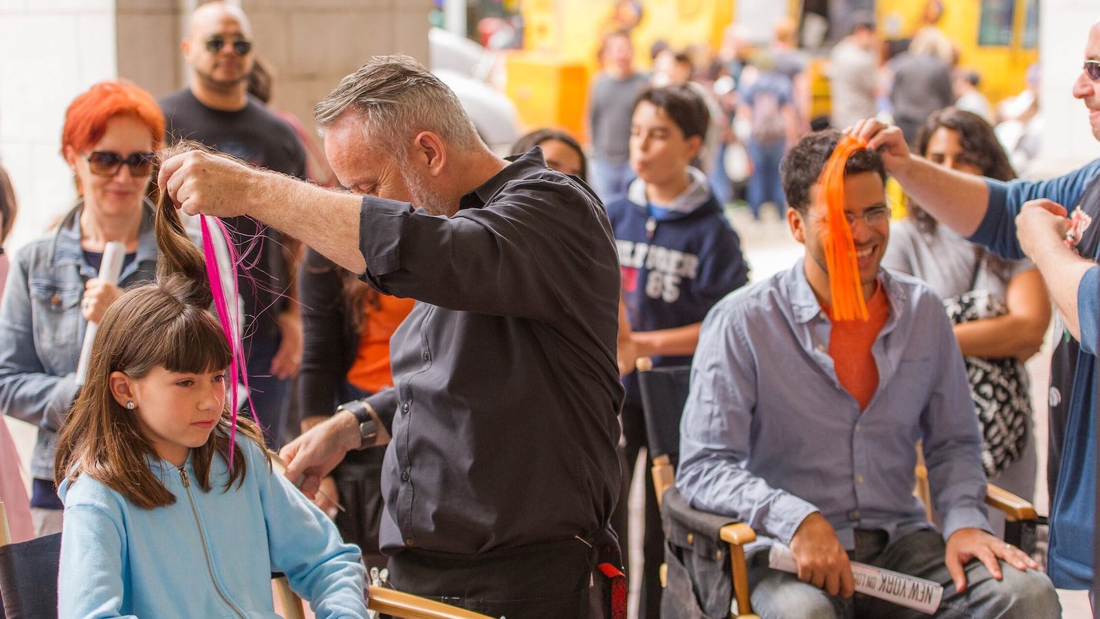 A young girl and older man sit in director folding chairs while men standing next to them hold colorful tassels up to their heads, as people surrounding them watch.