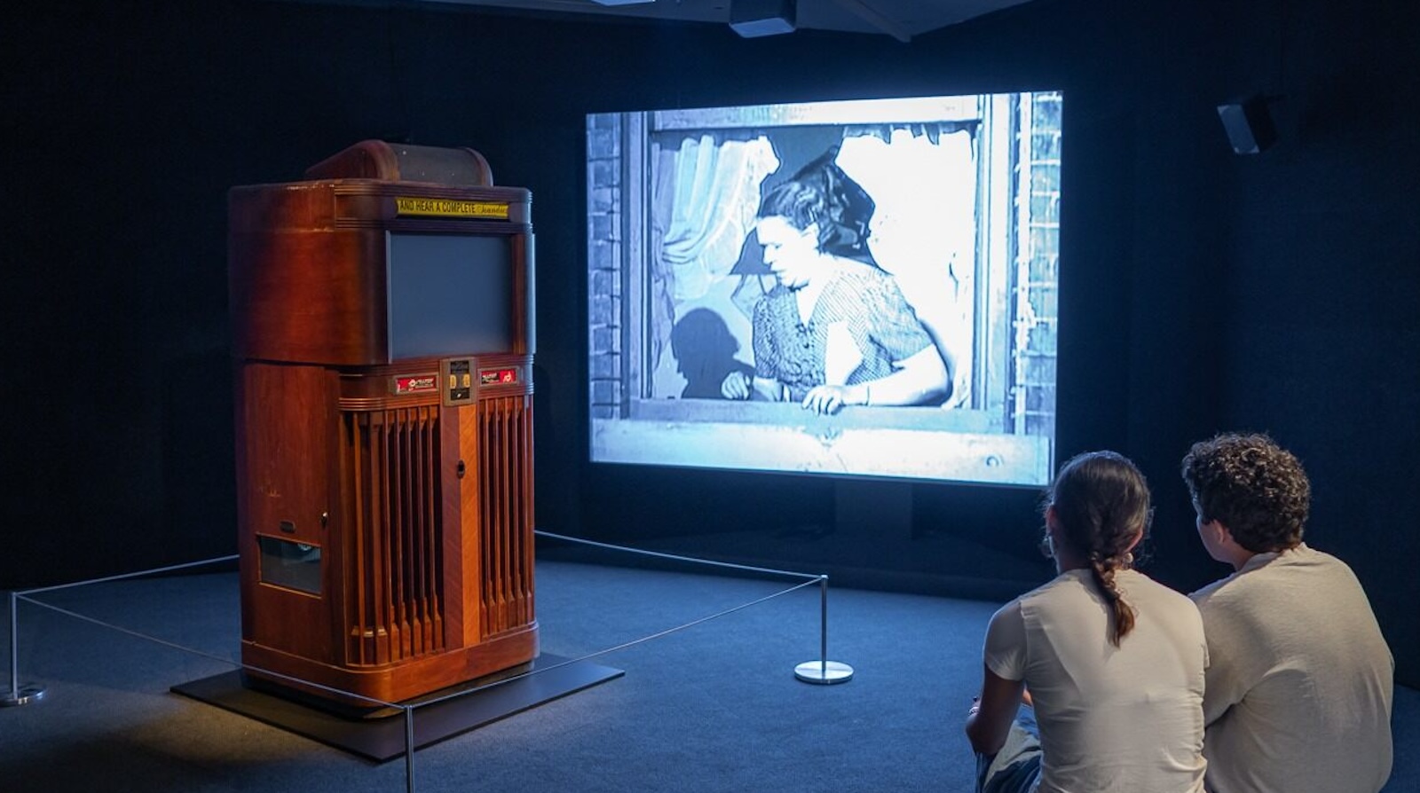 A Panoram machine, which looks like a wooden jukebox with a screen, sits in a Museum gallery, while on the wall behind it a black and white image of a woman looking out of a window is projected on the wall. Two people sit and watch, their backs to us.