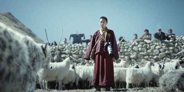 A Tibetan monk in a red robe stands amid a flock of sheep in a standoff with a white snow leopard facing him, seen slightly on the left of the frame