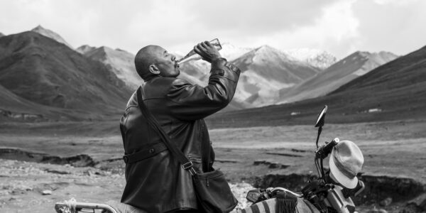 Against a mountainous backdrop a man in leather jacket drinks from a glass bottle as he sits on his motorcycle.
