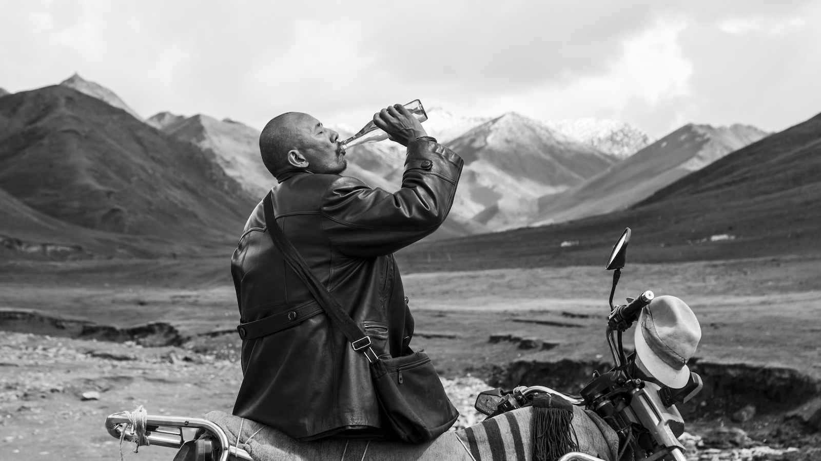 Against a mountainous backdrop a man in leather jacket drinks from a glass bottle as he sits on his motorcycle.