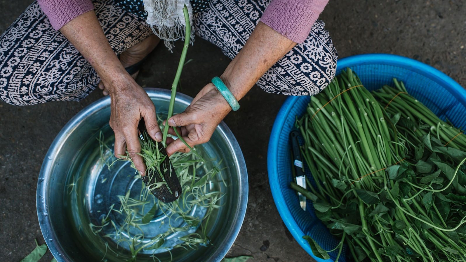 An overhead shot of slightly wrinkled hands shaving green vegetable stalks from one bowl into another.