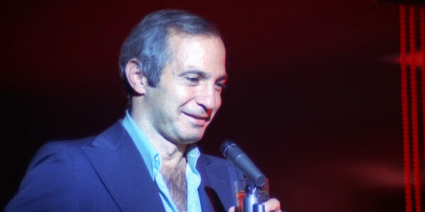 A man in blue suit, his shirt open to reveal chest hair, stands and smiles into a mic against a dark stage with red streaks on the right