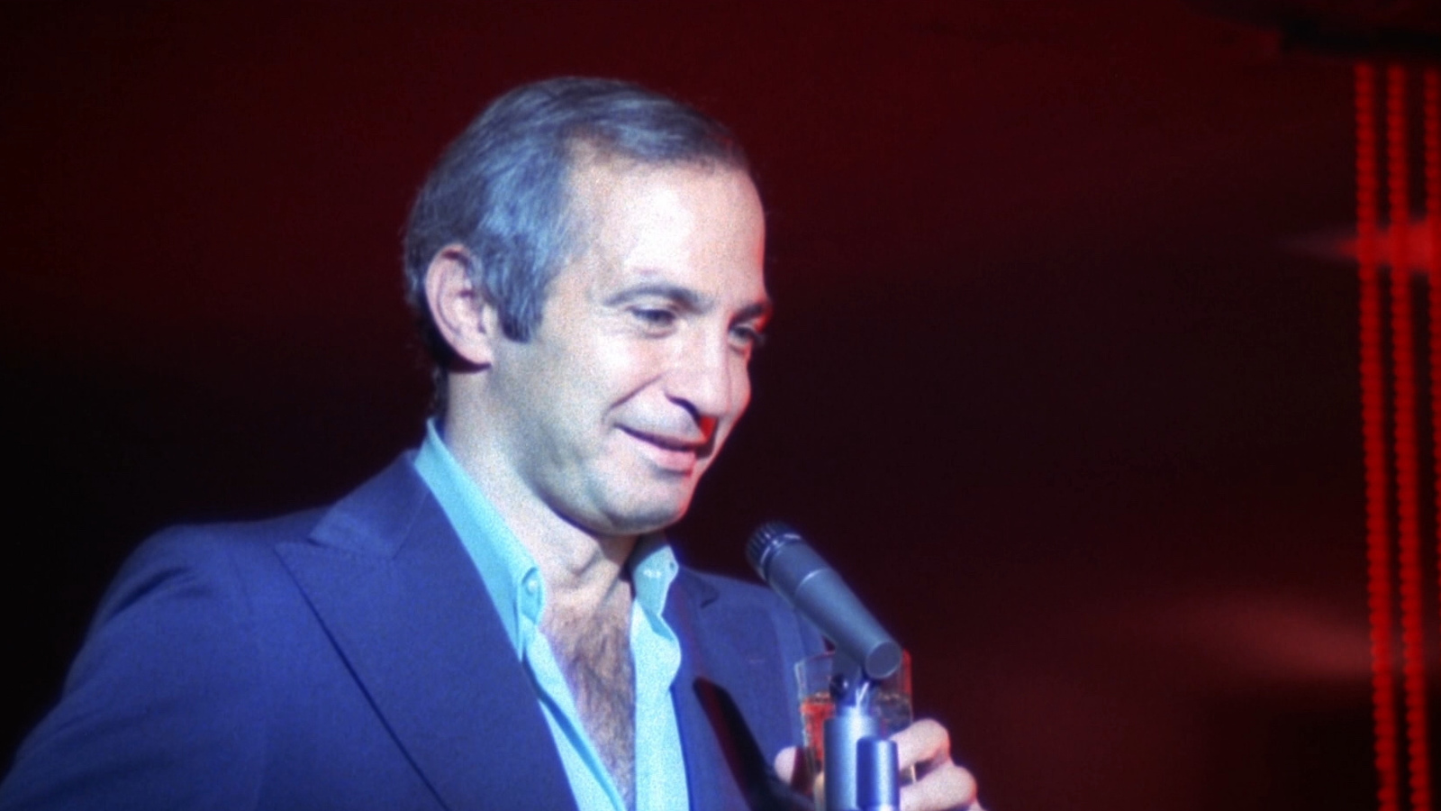 A man in blue suit, his shirt open to reveal chest hair, stands and smiles into a mic against a dark stage with red streaks on the right