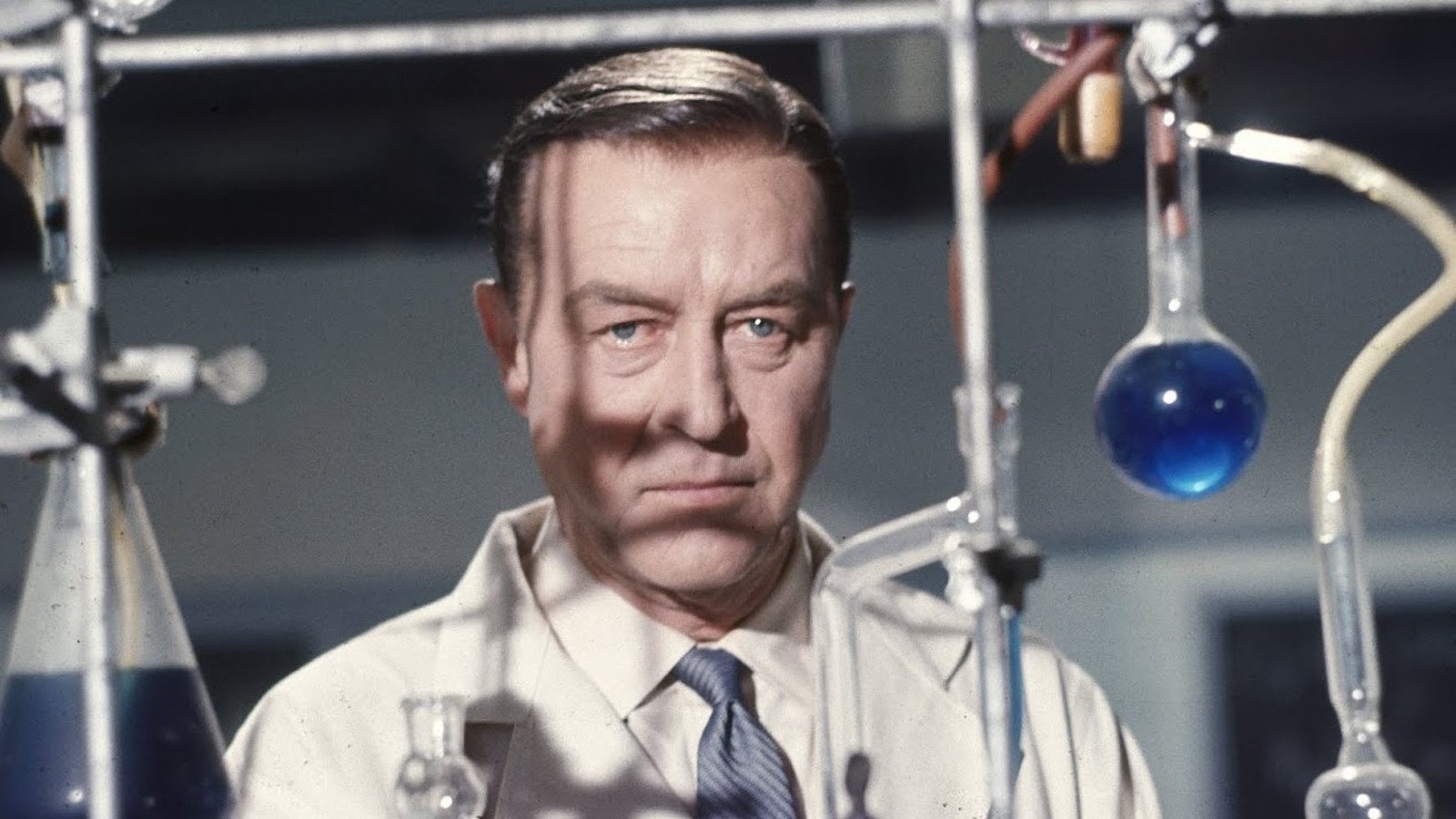 A man in a lab coat stands behind science lab equipment including test tubes, and looks off with a serious expression