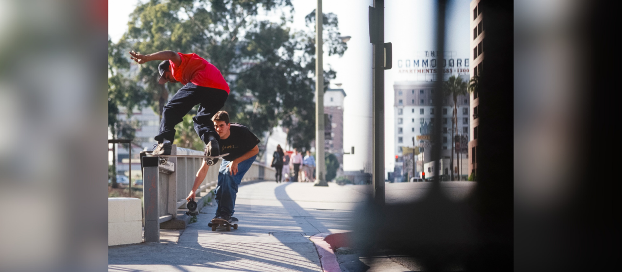 A skateboarder in a red T-shirt does a trick on a railing on a sidewalk while another skateboarder follow him with a camera held low to the ground.