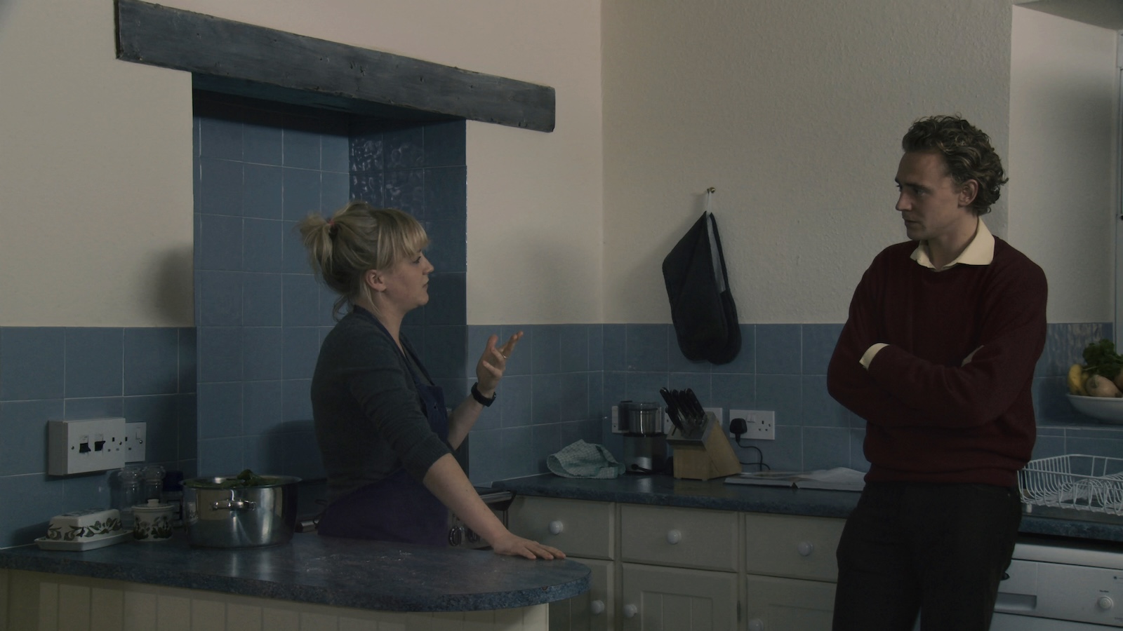 A man and woman stand in a chill blue kitchen talking; his arms are folded, she gestures with her left hand and the other leaning on counter