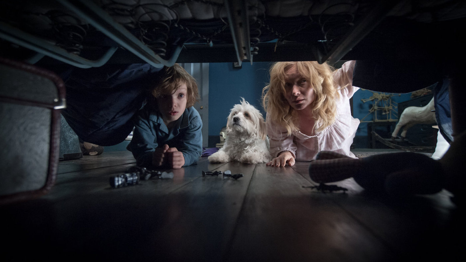 A little boy with tousled hair, a white puppy, and a blonde woman in a nightgown peer under a bed in a dark bedroom
