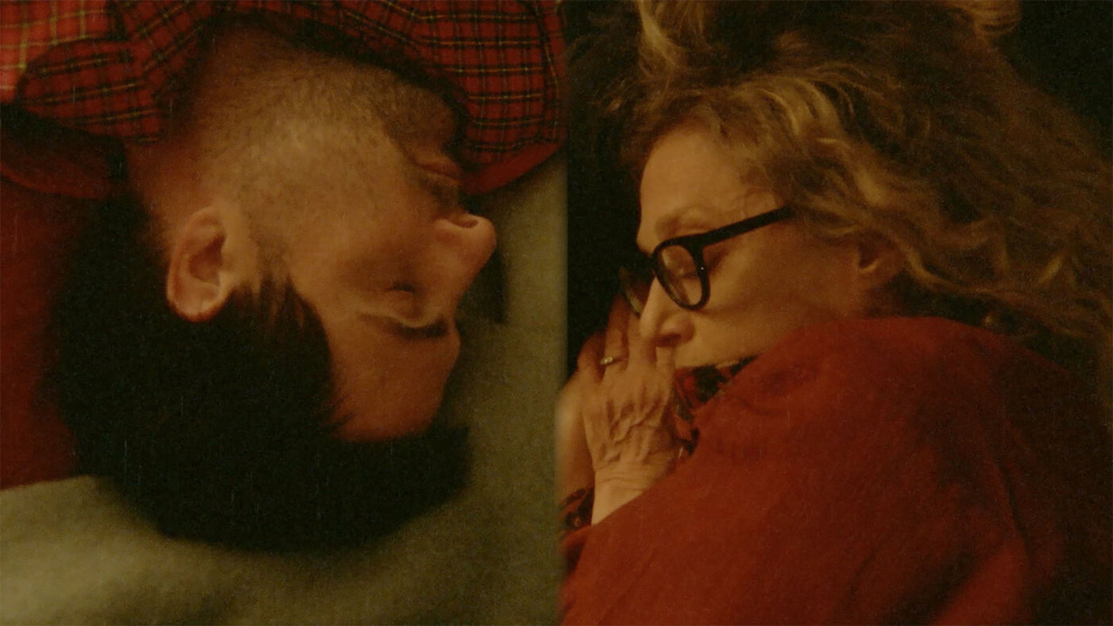 A split screen of a blonde woman in glasses and a dark haired man sleeping in separate beds facing each other, with the man upside down on the left.