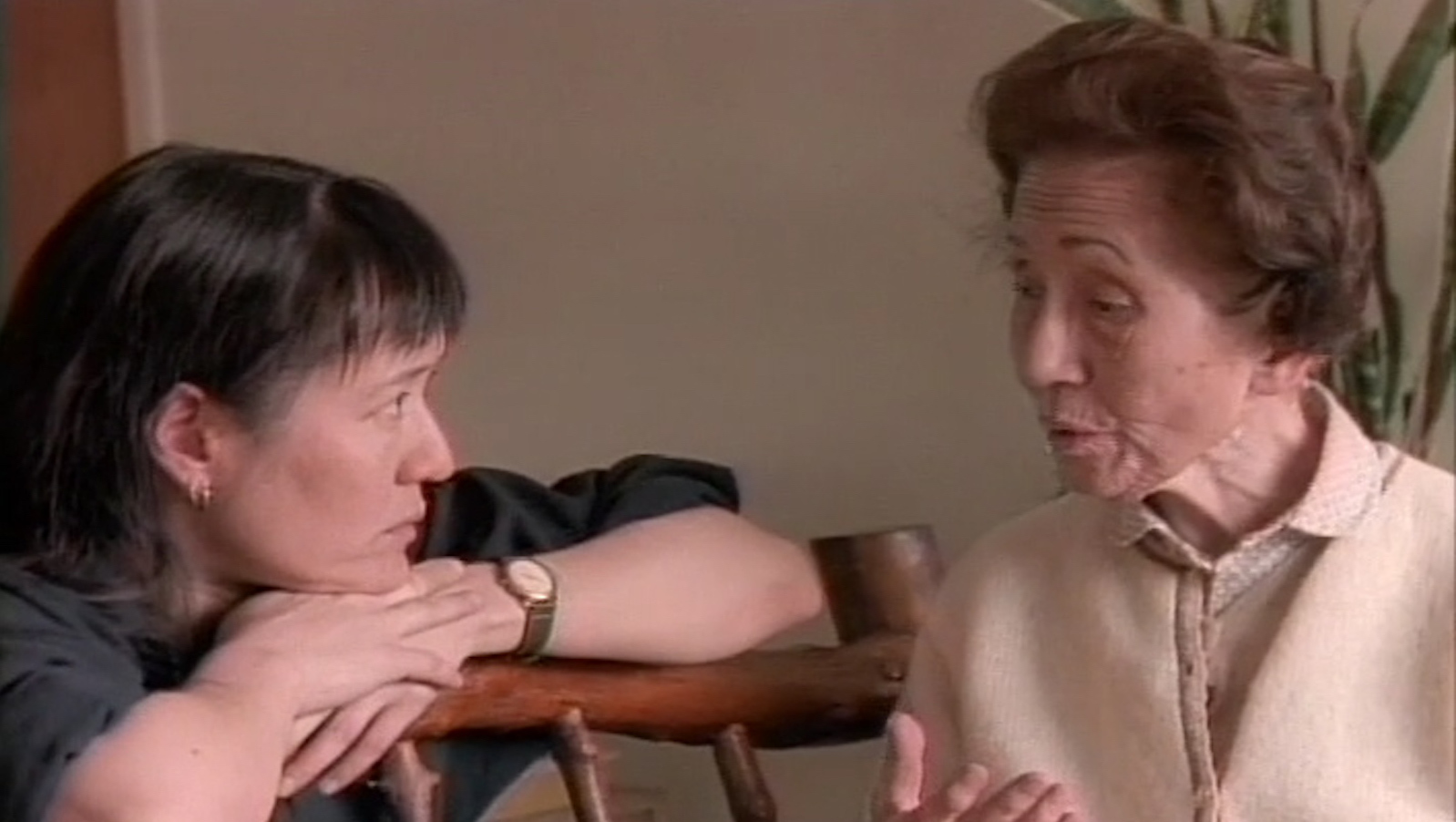 A woman rests her hands on the back of a chair while listening to an older woman talk next to her