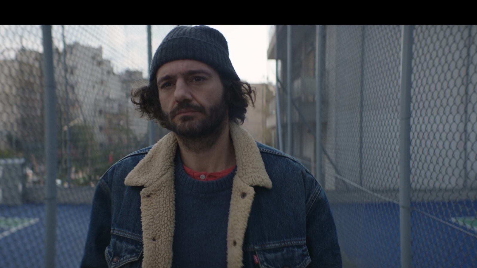 A man in a winter knit hat and jean jacket stands next to a chainlink fence in a city looking forlorn